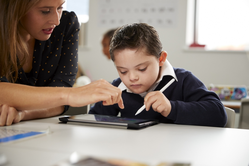 Young female teacher working with a Down syndrome schoolboy sitting at desk using a tablet computer